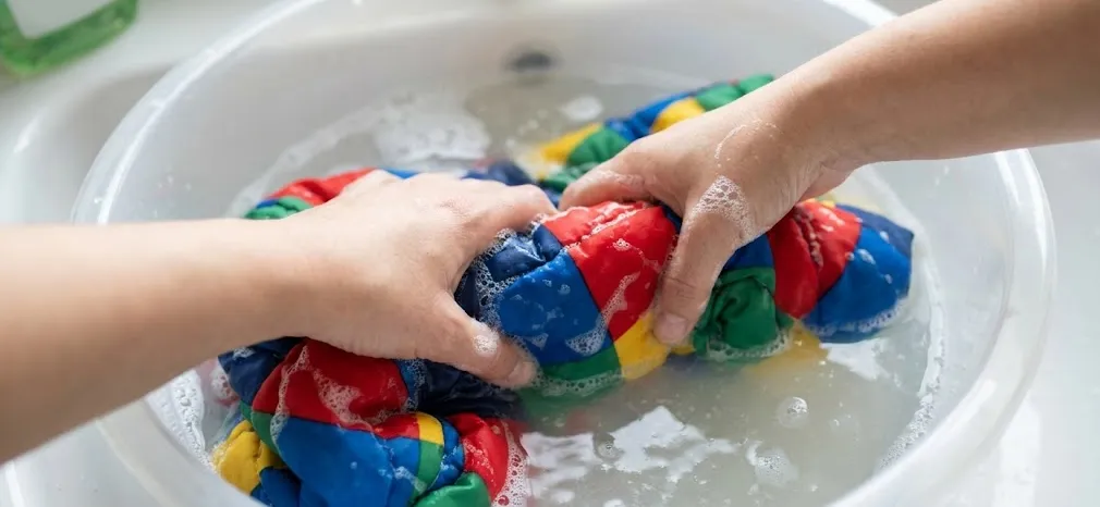 따뜻한 비눗물이 담긴 세탁조에 아이의 밝은색 패딩 재킷을 부드럽게 담그고 있는 손.